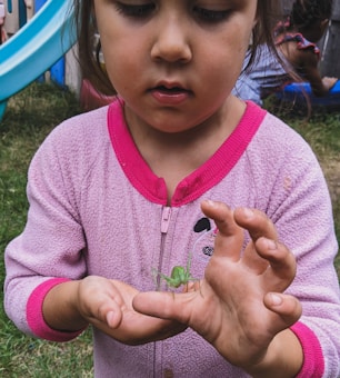 A young child wearing a pink outfit is focused on holding a green grasshopper gently in their hand. In the background, there is a brightly colored slide and another child playing.