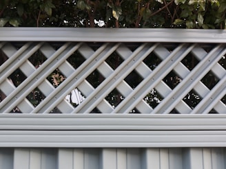 A lattice fence with a crisscross pattern made of evenly spaced horizontal and diagonal metal strips. The fence is painted in a muted light gray color and stands in front of dense green foliage, creating a contrast between the structured fence and the natural leaves.