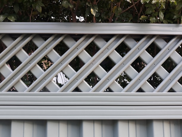 A lattice fence with a crisscross pattern made of evenly spaced horizontal and diagonal metal strips. The fence is painted in a muted light gray color and stands in front of dense green foliage, creating a contrast between the structured fence and the natural leaves.