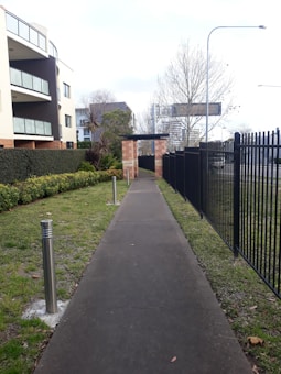 A paved walkway leads between a manicured grassy area with bushes on one side and a black metal fence on the other. The path is flanked by modern silver bollards. In the background, there's a large brick apartment building with glass balconies and some bare trees. A billboard and streetlights are visible in the distance.