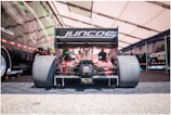 A racing car is positioned in a pit area with large rear tires and a prominent rear wing featuring the logo 'Juncos Racing'. The car is under a tent with a patterned floor, and there are people in the background working around the vehicle.