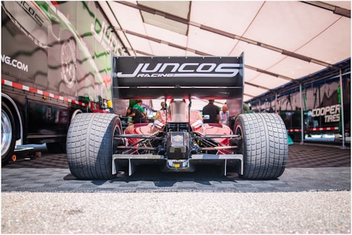 A racing car is positioned in a pit area with large rear tires and a prominent rear wing featuring the logo 'Juncos Racing'. The car is under a tent with a patterned floor, and there are people in the background working around the vehicle.