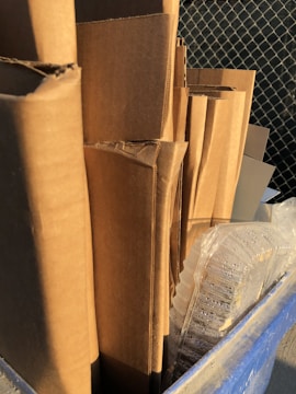 A stack of neatly arranged cardboard boxes sits in a recycling bin. The boxes are standing upright, and next to them is a clear plastic container with droplets of water on its surface. The bin is blue, and a wire fence is visible in the background.