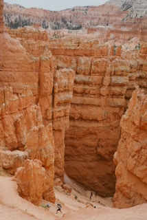 Steep canyon walls with distinct hoodoo formations dominate the landscape. Hikers are seen making their way along the sandy path at the base, providing a sense of scale to the towering rock structures. The scene is characterized by contrasting textures of rugged stone and sparse vegetation in a dry, arid environment.