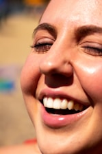 A close-up of a person's face with a joyful expression, eyes closed and mouth open in a smile, showcasing shining white teeth. The skin appears smooth and slightly sunlit.
