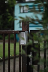 A close-up of a metallic gate with an intercom or doorbell system attached. The gate is slightly ajar and surrounded by greenery, with a blurred view of a blue and white building in the background. The mood is calm and tranquil, suggesting a residential area.