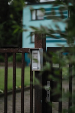 A close-up of a metallic gate with an intercom or doorbell system attached. The gate is slightly ajar and surrounded by greenery, with a blurred view of a blue and white building in the background. The mood is calm and tranquil, suggesting a residential area.