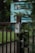 A close-up of a metallic gate with an intercom or doorbell system attached. The gate is slightly ajar and surrounded by greenery, with a blurred view of a blue and white building in the background. The mood is calm and tranquil, suggesting a residential area.