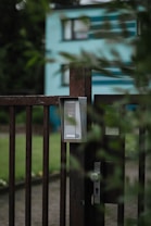 A close-up of a metallic gate with an intercom or doorbell system attached. The gate is slightly ajar and surrounded by greenery, with a blurred view of a blue and white building in the background. The mood is calm and tranquil, suggesting a residential area.