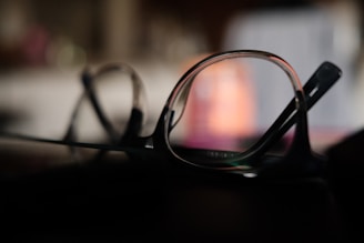 A close-up view of eyeglasses lying on a dark surface, with one lens in focus and the rest of the frame blurred. The background appears soft and unfocused, displaying subtle variations in color from dark to light shades.