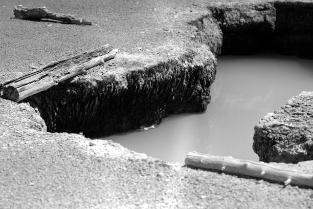 A black and white photograph featuring a large pothole filled with water surrounded by rough terrain. Several wooden logs lie scattered around the area, providing a rustic feel. The texture of the rough surface contrasts with the smoothness of the water.