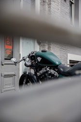 A green motorcycle is parked in front of a building with a gray brick wall and a door that has a sign indicating 'Coming Soon'. The motorcycle is partially obscured by a horizontal barrier in the foreground, adding depth to the composition.