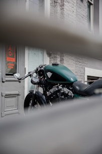 A green motorcycle is parked in front of a building with a gray brick wall and a door that has a sign indicating 'Coming Soon'. The motorcycle is partially obscured by a horizontal barrier in the foreground, adding depth to the composition.