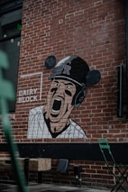 A mural painted on a brick wall features a baseball player wearing a helmet and striped uniform, shouting or cheering with an intense expression. The words 'DAIRY BLOCK' and 'LET'S GO ROCKIES!' are also visible. Below the mural are some green metallic chairs and wooden blocks arranged as seats outdoors.