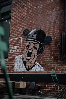A mural painted on a brick wall features a baseball player wearing a helmet and striped uniform, shouting or cheering with an intense expression. The words 'DAIRY BLOCK' and 'LET'S GO ROCKIES!' are also visible. Below the mural are some green metallic chairs and wooden blocks arranged as seats outdoors.
