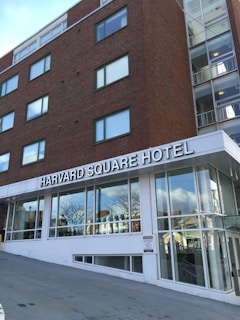 A multistory brick building with large windows featuring a sign for Harvard Square Hotel. The building is adjacent to a modern glass entrance, and the surroundings include other structures reflected in the windows.