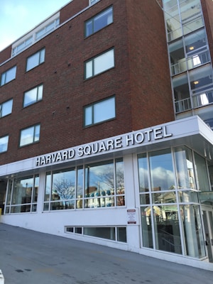 A multistory brick building with large windows featuring a sign for Harvard Square Hotel. The building is adjacent to a modern glass entrance, and the surroundings include other structures reflected in the windows.