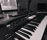 A monochrome image of a music production setup featuring a laptop displaying music software and a MIDI keyboard with various controls and buttons. The setup suggests a focus on electronic music production.