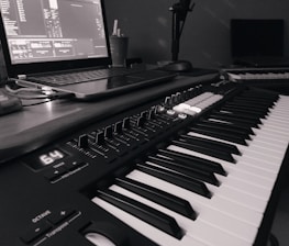 A monochrome image of a music production setup featuring a laptop displaying music software and a MIDI keyboard with various controls and buttons. The setup suggests a focus on electronic music production.