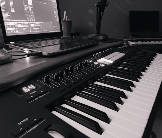 A monochrome image of a music production setup featuring a laptop displaying music software and a MIDI keyboard with various controls and buttons. The setup suggests a focus on electronic music production.