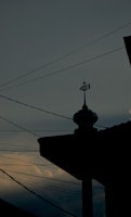 A silhouette of a mosque's rooftop is adorned with an Islamic symbol, set against a dusky sky with scattered clouds. Power lines stretch across the scene, intersecting with the darkened outline of the building.
