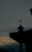 A silhouette of a mosque's rooftop is adorned with an Islamic symbol, set against a dusky sky with scattered clouds. Power lines stretch across the scene, intersecting with the darkened outline of the building.