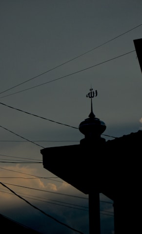 A silhouette of a mosque's rooftop is adorned with an Islamic symbol, set against a dusky sky with scattered clouds. Power lines stretch across the scene, intersecting with the darkened outline of the building.