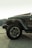 A partial view of a Jeep Wrangler Sahara model, showcasing its front tire and portion of the passenger side. The vehicle is parked on a concrete surface with a backdrop of shipping containers. The light in the scene is soft, suggesting either early morning or late afternoon.