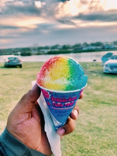 A hand holds a colorful snow cone with a red, yellow, green, and blue gradient, set against a backdrop of a grassy field and a cloudy sky by a body of water. Cars are parked in the distance.