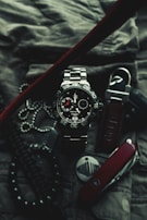 A close-up of a sophisticated wristwatch with a metallic band and black dial, featuring white and red accents. The watch is surrounded by various accessories, including a Swiss army knife, a set of dog tags, and a textured fabric background, suggesting a rugged and adventurous theme.