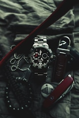 A close-up of a sophisticated wristwatch with a metallic band and black dial, featuring white and red accents. The watch is surrounded by various accessories, including a Swiss army knife, a set of dog tags, and a textured fabric background, suggesting a rugged and adventurous theme.