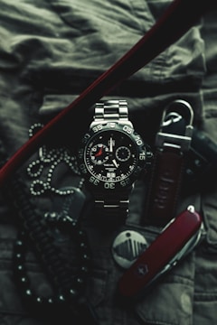 A close-up of a sophisticated wristwatch with a metallic band and black dial, featuring white and red accents. The watch is surrounded by various accessories, including a Swiss army knife, a set of dog tags, and a textured fabric background, suggesting a rugged and adventurous theme.
