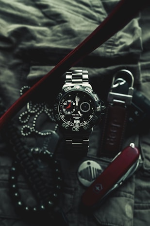 A close-up of a sophisticated wristwatch with a metallic band and black dial, featuring white and red accents. The watch is surrounded by various accessories, including a Swiss army knife, a set of dog tags, and a textured fabric background, suggesting a rugged and adventurous theme.