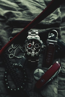 A close-up of a sophisticated wristwatch with a metallic band and black dial, featuring white and red accents. The watch is surrounded by various accessories, including a Swiss army knife, a set of dog tags, and a textured fabric background, suggesting a rugged and adventurous theme.