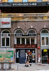 A city street with a brick building housing multiple businesses. At the top, there's a sign for a karaoke place with Korean characters. Below, there's a sign for a fitness center called Nimble Fitness and a venue named Barcade with a neon sign. A person wearing a mask walks past the entrance that displays various signs and posters. To the left, a phone booth is visible with an advertisement on it.