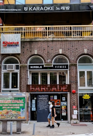 A city street with a brick building housing multiple businesses. At the top, there's a sign for a karaoke place with Korean characters. Below, there's a sign for a fitness center called Nimble Fitness and a venue named Barcade with a neon sign. A person wearing a mask walks past the entrance that displays various signs and posters. To the left, a phone booth is visible with an advertisement on it.