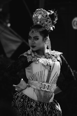 A performer dressed in elaborate traditional attire featuring an ornate headdress and detailed embroidery strikes a confident pose. The image is in black and white, enhancing the dramatic effect and focus on the intricate costume details.