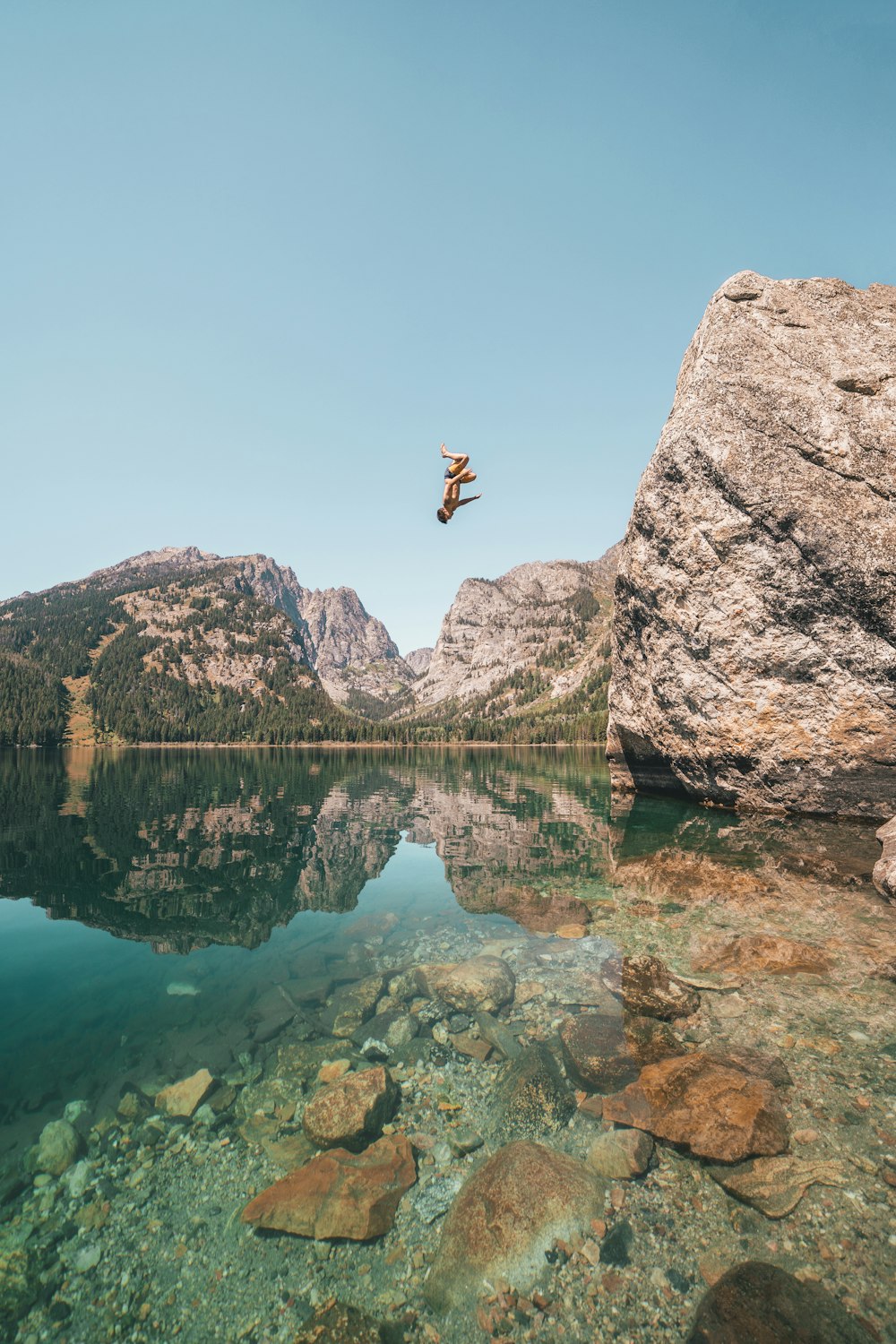 person jumping from rock formation to lake during daytime
