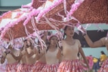 A group of performers is dressed in coordinated outfits featuring beige tops and colorful skirts, holding large umbrellas adorned with pink decorations. They appear to be participating in a parade or festival, moving in unison and displaying expressions of concentration and joy.