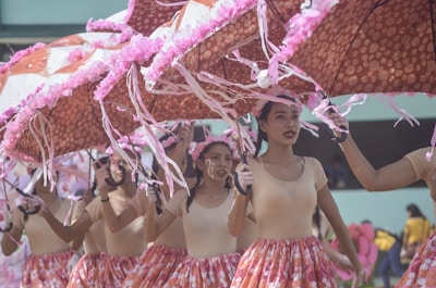 A group of performers is dressed in coordinated outfits featuring beige tops and colorful skirts, holding large umbrellas adorned with pink decorations. They appear to be participating in a parade or festival, moving in unison and displaying expressions of concentration and joy.
