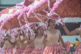 A group of performers is dressed in coordinated outfits featuring beige tops and colorful skirts, holding large umbrellas adorned with pink decorations. They appear to be participating in a parade or festival, moving in unison and displaying expressions of concentration and joy.