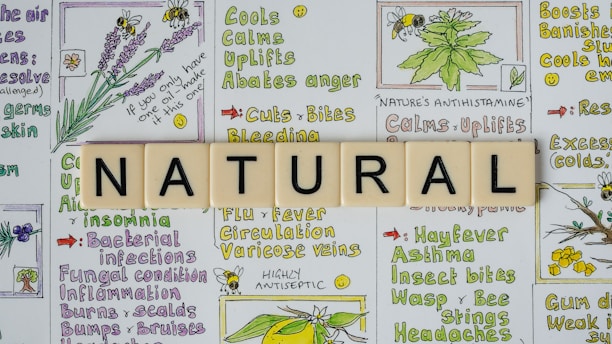 The image features the word 'NATURAL' spelled out with Scrabble tiles placed on top of a background filled with colorful illustrations and text about herbal remedies. The background contains drawings of plants like lavender and other herbs, alongside descriptions of their benefits, such as calming, uplifting, and treating various ailments.