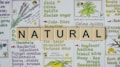 The image features the word 'NATURAL' spelled out with Scrabble tiles placed on top of a background filled with colorful illustrations and text about herbal remedies. The background contains drawings of plants like lavender and other herbs, alongside descriptions of their benefits, such as calming, uplifting, and treating various ailments.