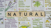 The image features the word 'NATURAL' spelled out with Scrabble tiles placed on top of a background filled with colorful illustrations and text about herbal remedies. The background contains drawings of plants like lavender and other herbs, alongside descriptions of their benefits, such as calming, uplifting, and treating various ailments.