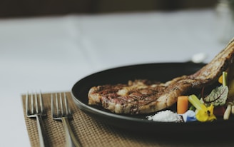A black plate with a grilled ribeye steak garnished with colorful vegetables and a small pile of coarse salt. The setup includes two forks placed neatly on a woven placemat.