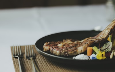 A black plate with a grilled ribeye steak garnished with colorful vegetables and a small pile of coarse salt. The setup includes two forks placed neatly on a woven placemat.