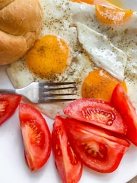 A breakfast setting featuring two sunny-side-up eggs seasoned with black pepper, accompanied by a sliced bread roll. Fresh tomato slices are arranged beside the eggs, enhancing the vibrant color and inviting presentation. A metal fork lies nearby, suggesting a ready-to-eat meal.