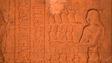 A detailed stone carving features an ancient Egyptian figure with braided hair, adorned with jewelry, extending their hands towards a series of ornate vessels. Hieroglyphics run vertically alongside the figure, intricately etched into the stone surface.