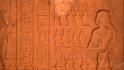 A detailed stone carving features an ancient Egyptian figure with braided hair, adorned with jewelry, extending their hands towards a series of ornate vessels. Hieroglyphics run vertically alongside the figure, intricately etched into the stone surface.