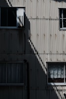 A corrugated metal wall with a retro-industrial appearance, featuring several rectangular windows. The wall shows a distinct angular shadow casting across its surface, enhancing the texture and creating a play of light and dark.